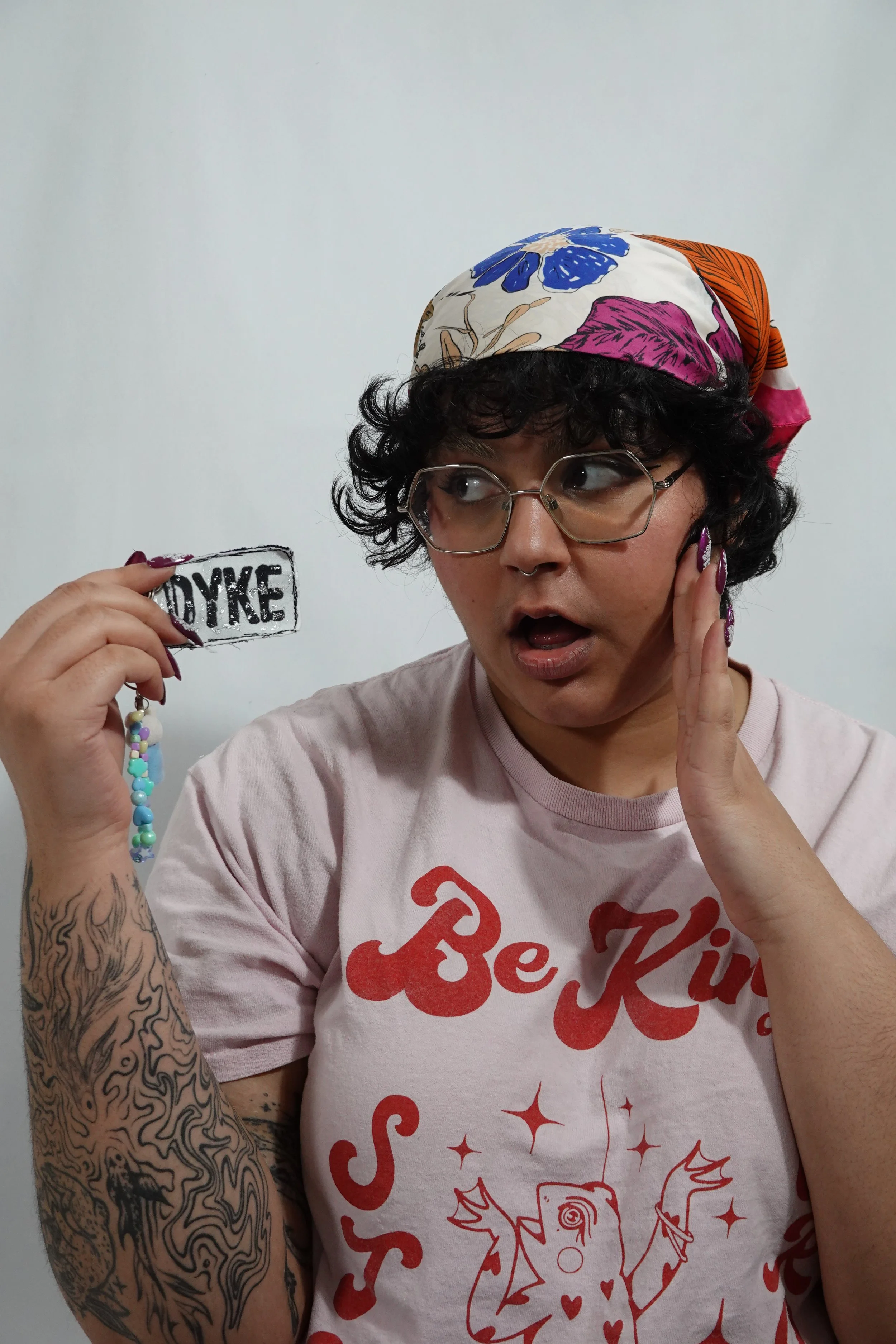 Femme with glasses holding a sign that says 'DYKE', wearing a pink T-shirt with red text and a graphic, and a colorful headscarf, appearing surprised or shocked.