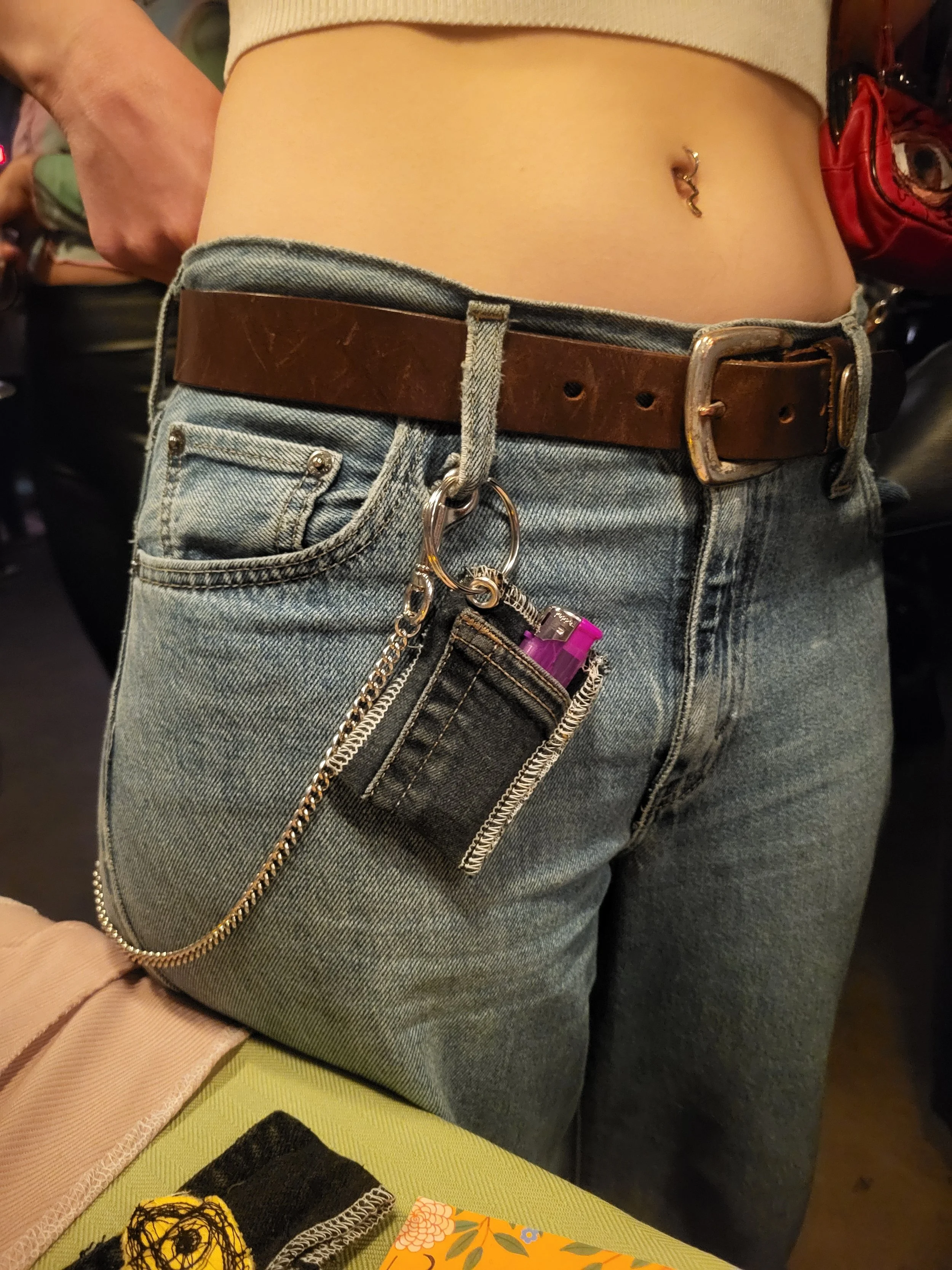 Close-up of a person's waist wearing distressed jeans, a brown belt with a keychain holding a small black pouch with a zipper and a pink lighter inside, and a small belly button piercing.