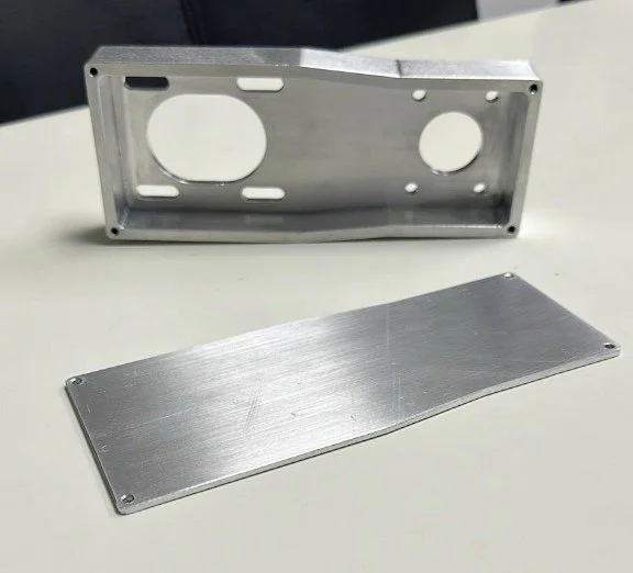 Two silver-colored metal pieces, one with cutouts and the other flat and rectangular, placed on a white surface.