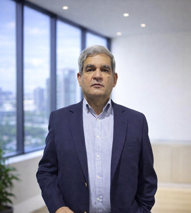 A man with gray hair wearing a dark blue blazer and light blue striped shirt standing in a modern office with large windows showing a cityscape outside.