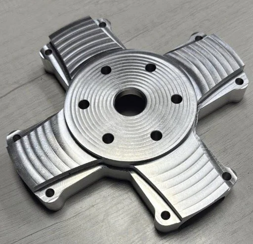 A metallic, circular mechanical part with four extended arms, each with curved grooves and holes, resting on a light-colored wooden surface.