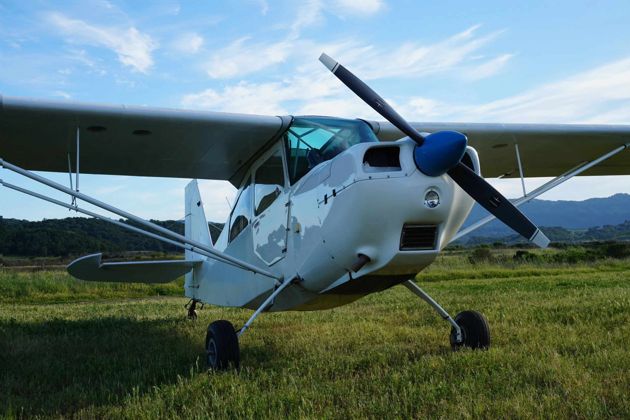 White tail wheel training airplane at Gnoss Field in Novato, with high wings and a propeller, parked on green grass with a backdrop of hills and a partly cloudy sky.