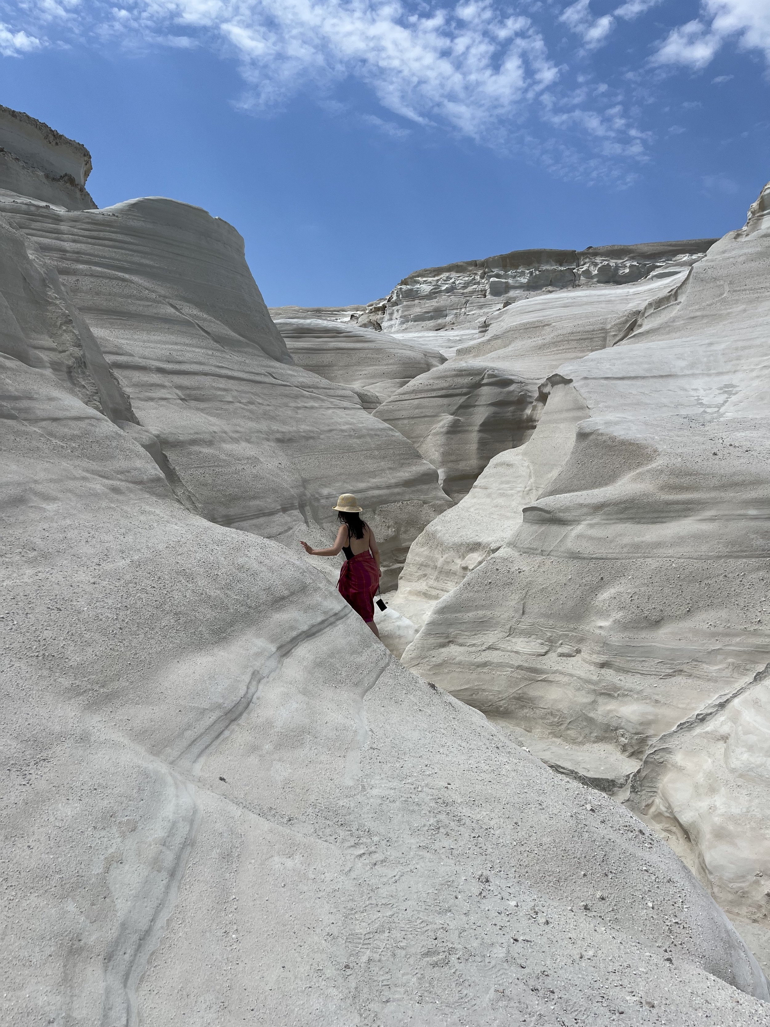 A woman wearing a hat and a red dress or sarong is walking through a narrow canyon with white and gray layered rock walls under a blue sky with some clouds.