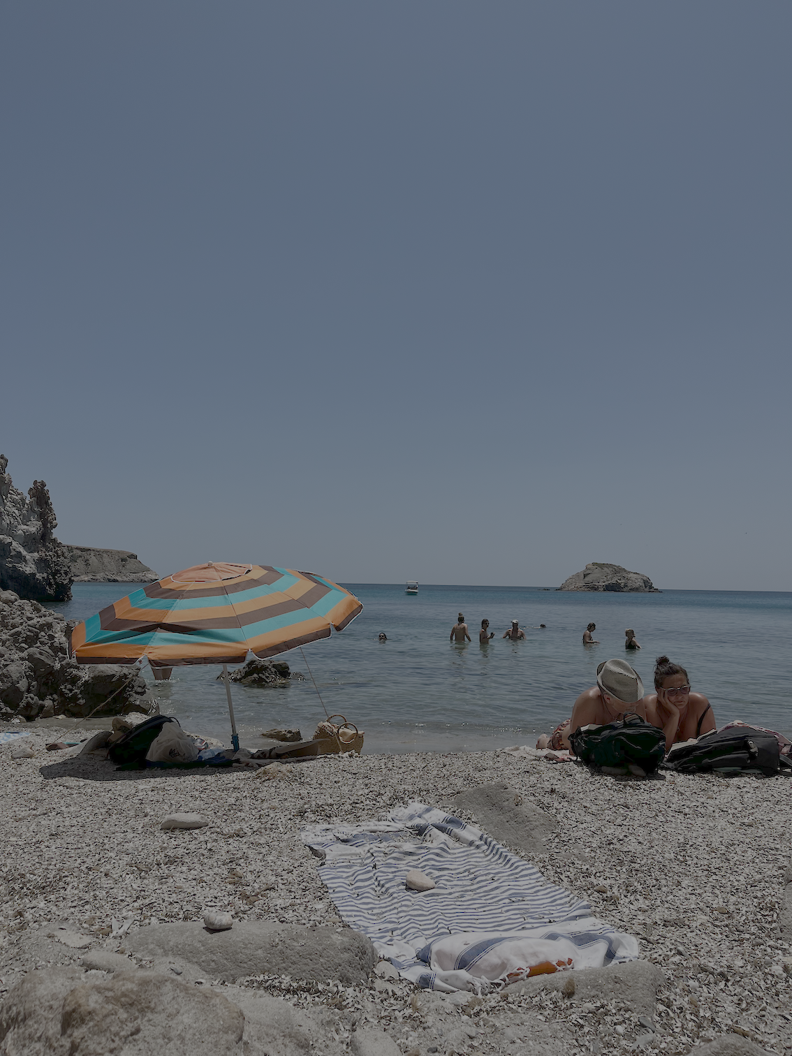 Beach scene with a colorful striped umbrella, towels, and rocks in the foreground. Several people are swimming and relaxing in the water, with rocks and small islands in the background under a clear blue sky.