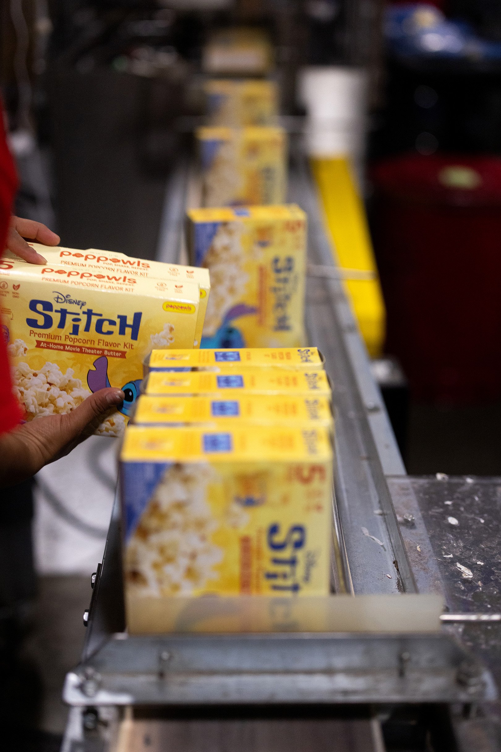 A person holding a Disney Stitch popcorn flavor kit in a factory. The kit is yellow with blue and red text, and is being held near other similar popcorn kits on a conveyor belt.