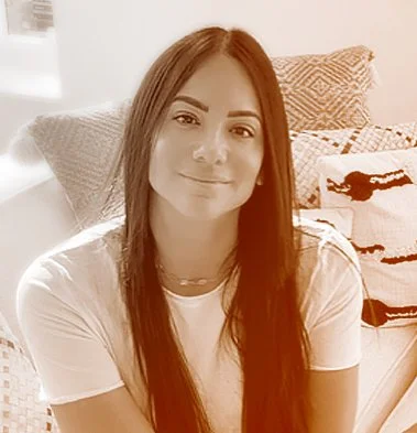 A young woman with long brown hair, smiling, sitting in a room with a bed and pillows in the background.