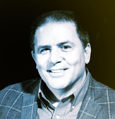 Portrait of a man with short curly hair wearing a suit jacket and collared shirt, smiling at the camera.