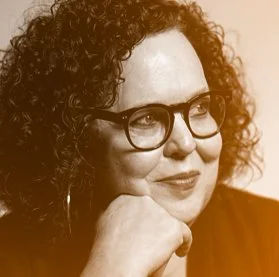 A woman with curly hair and glasses resting her chin on her hand, looking away thoughtfully.