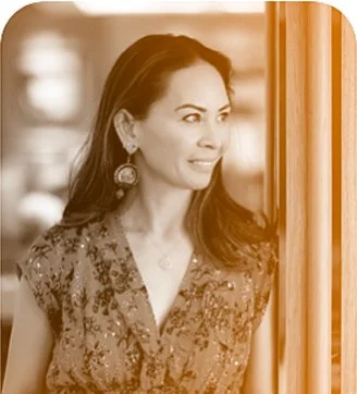 A woman with long dark hair wearing a patterned top and earrings, looking out of a window.