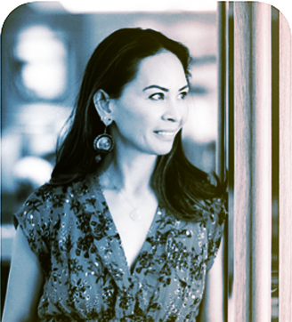 A woman with long dark hair wearing a patterned top and earrings, looking out of a window.