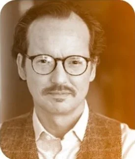 A portrait of a man with glasses, a mustache, and curly hair, wearing a collared shirt and sweater vest.