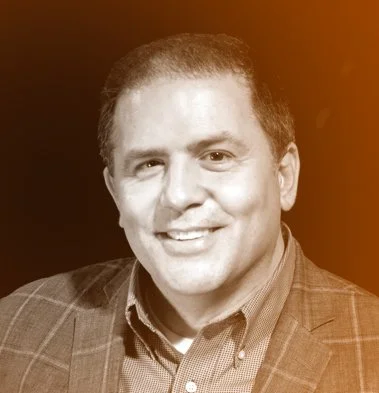 Portrait of a man with short curly hair wearing a suit jacket and collared shirt, smiling at the camera.