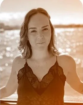 A woman with wavy hair standing outdoors by water at sunset, wearing a black sleeveless top with lace details.