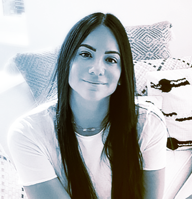 A young woman with long brown hair, smiling, sitting in a room with a bed and pillows in the background.