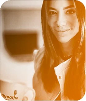A woman with long dark hair smiling at the camera, holding a white mug with the word 'breathe' on it