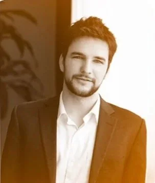A young man with dark hair and a beard, wearing a suit and dress shirt, standing indoors near a window.