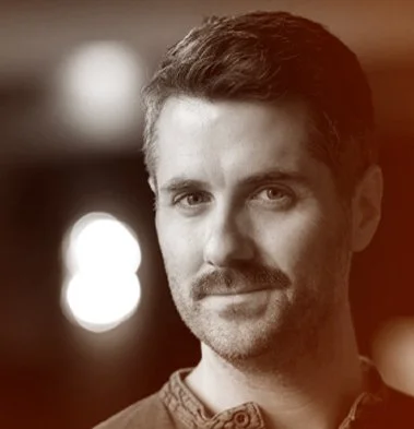 Black-and-white portrait of a man with short hair and a mustache, looking directly at the camera with a slight smile.