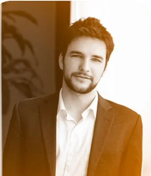 A young man with dark hair and a beard, wearing a suit and white shirt, standing indoors near a window with a plant in the background.