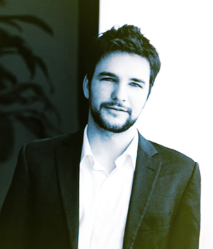 A young man with dark hair and a beard, wearing a suit and dress shirt, standing indoors near a window.