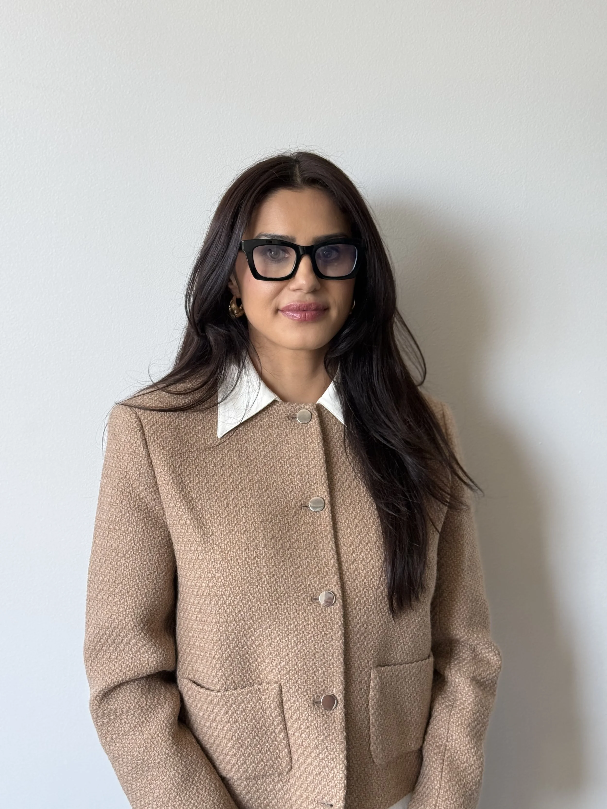 A woman with long dark hair wearing large black glasses, gold hoop earrings, a white collared shirt, and a beige textured blazer standing in front of a plain white wall.