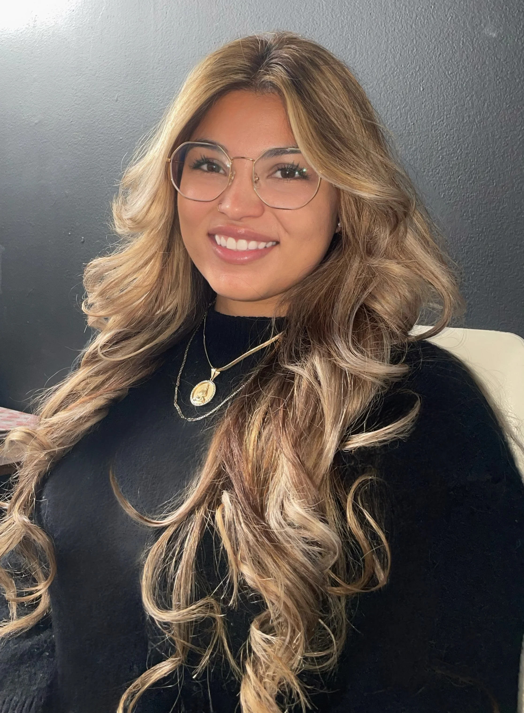 A young woman with long, wavy blonde hair and glasses, smiling, wearing a black top and layered gold necklaces, sitting against a dark wall.