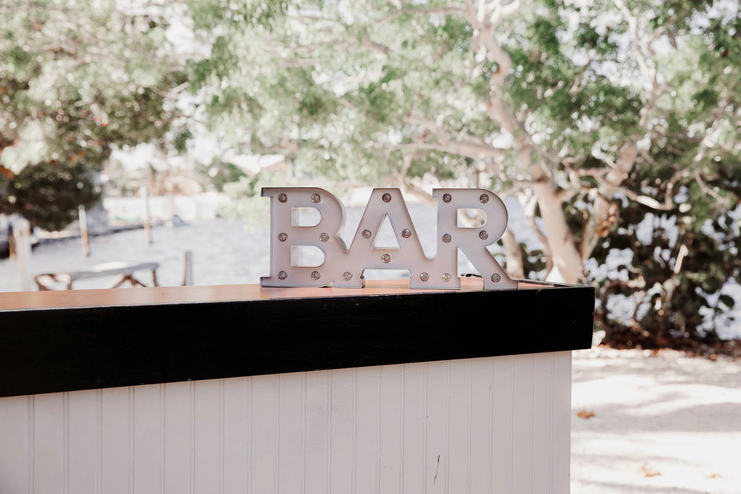 Decorative sign spelling 'BAR' with large metallic letters and light bulbs, placed on a counter outdoors with trees in the background.