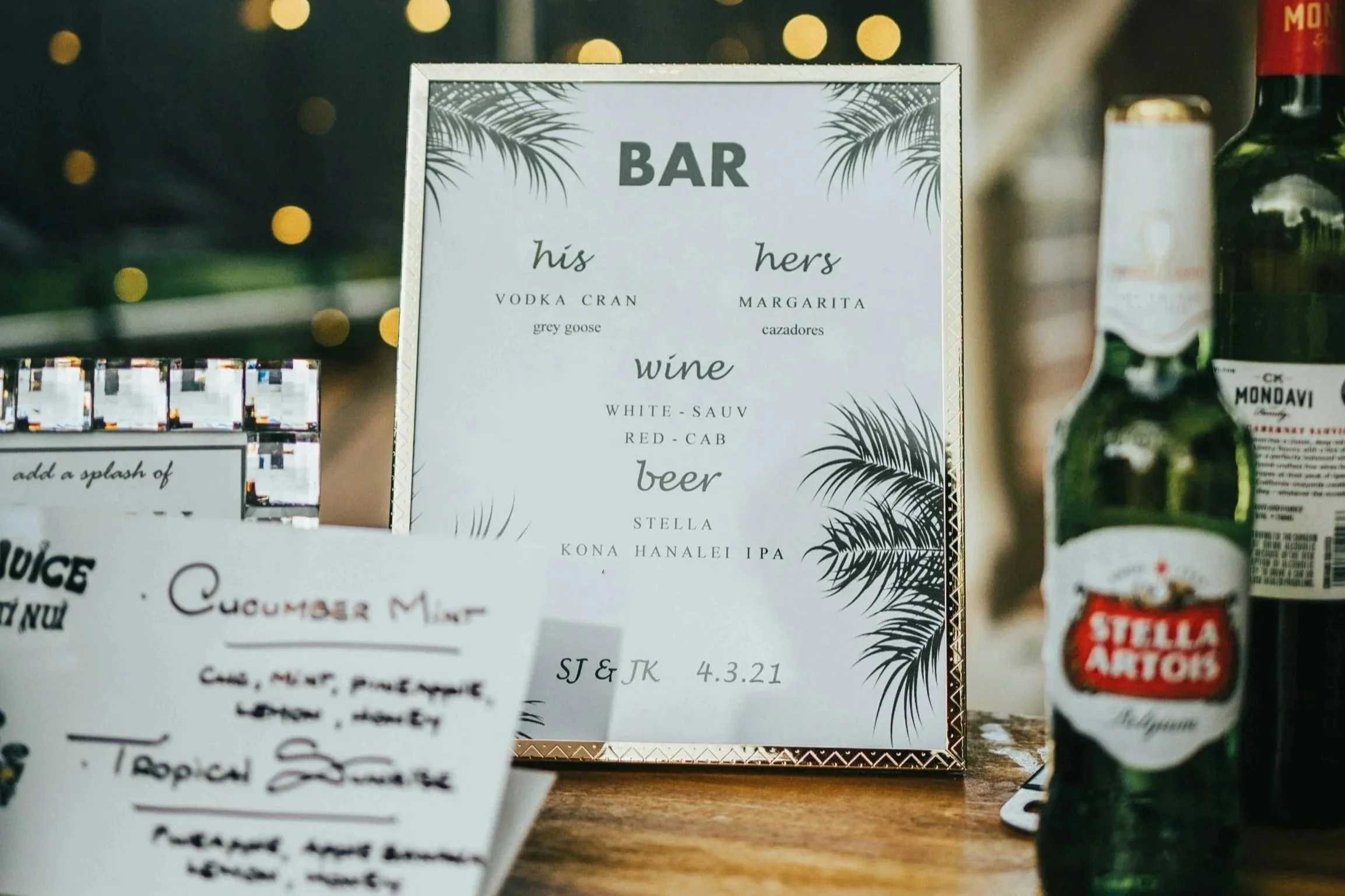 A menu sign at a bar featuring drinks for him and her, including vodka cranberry and margarita, with wine options for white-sauv, red-cab, and beer Stella, Kona, Hanalei IPA, and others. The sign has a tropical design with palm leaves.