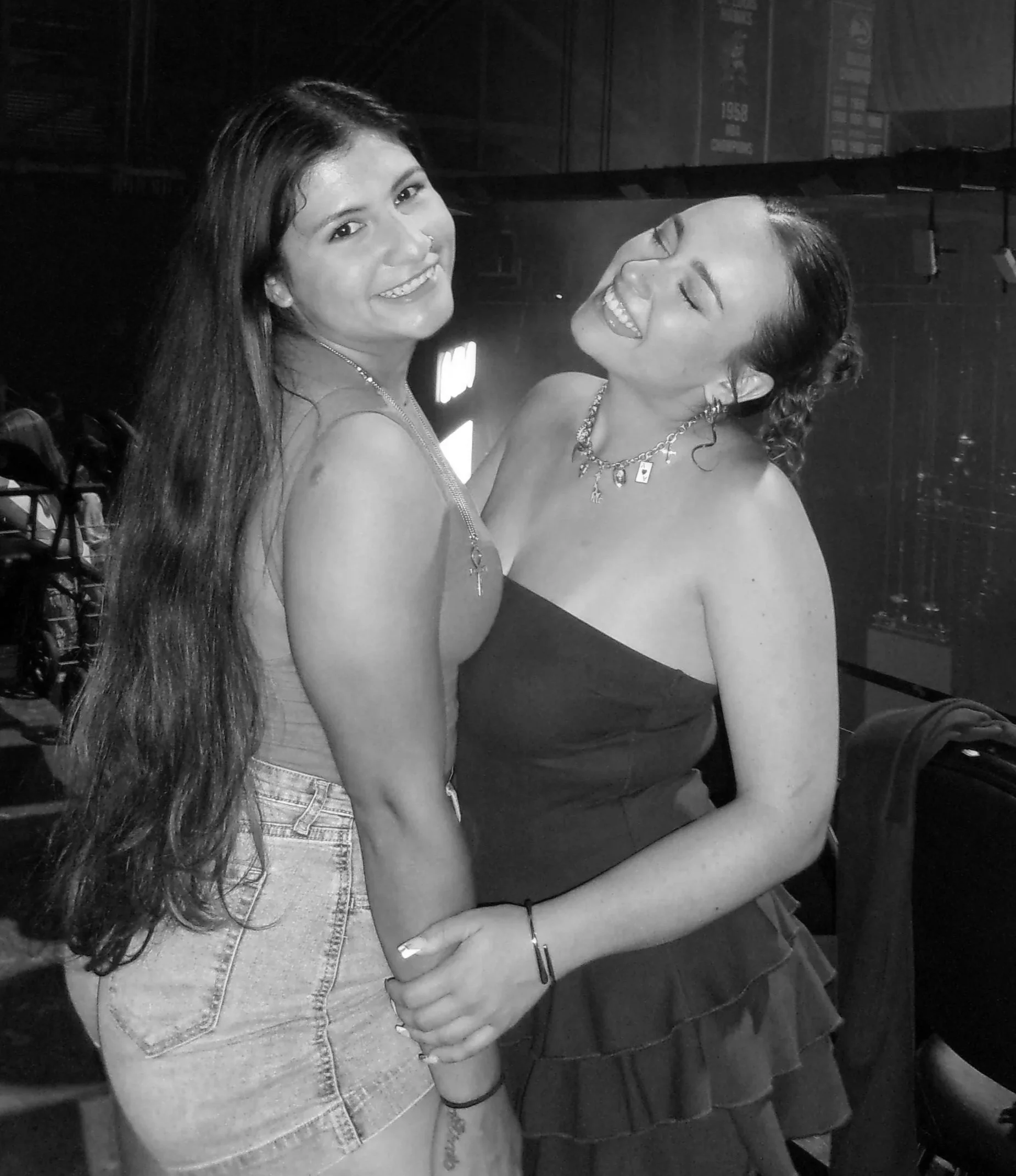 Two young women smiling and posing closely together in a black and white photo, one wearing a denim jacket and the other in a strapless top with layered ruffles, both wearing jewelry including necklaces and earrings.