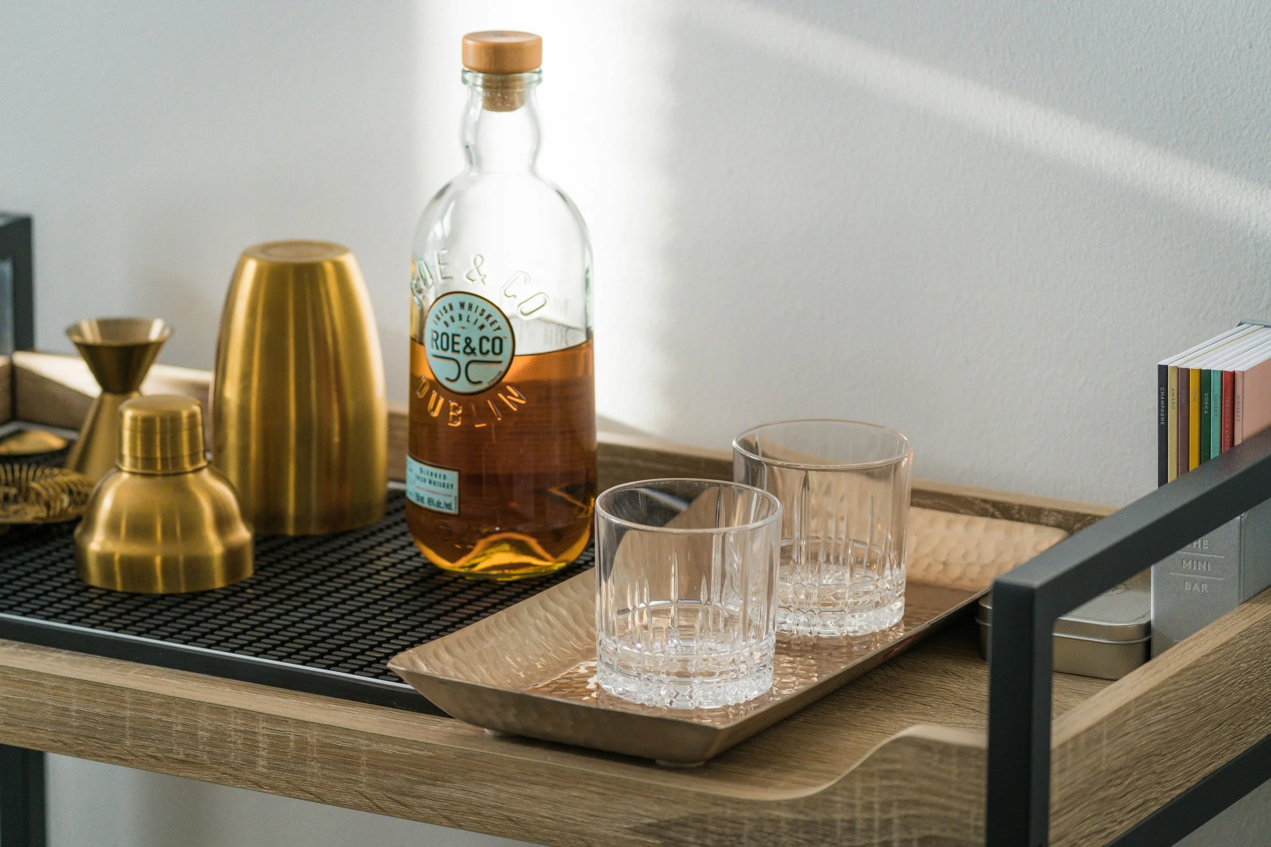 A bar cart with a bottle of Roe & Co Irish whiskey, gold cocktail shakers, a gold jigger, and empty glasses on a black tray and a gold tray. There are also books on the right side.