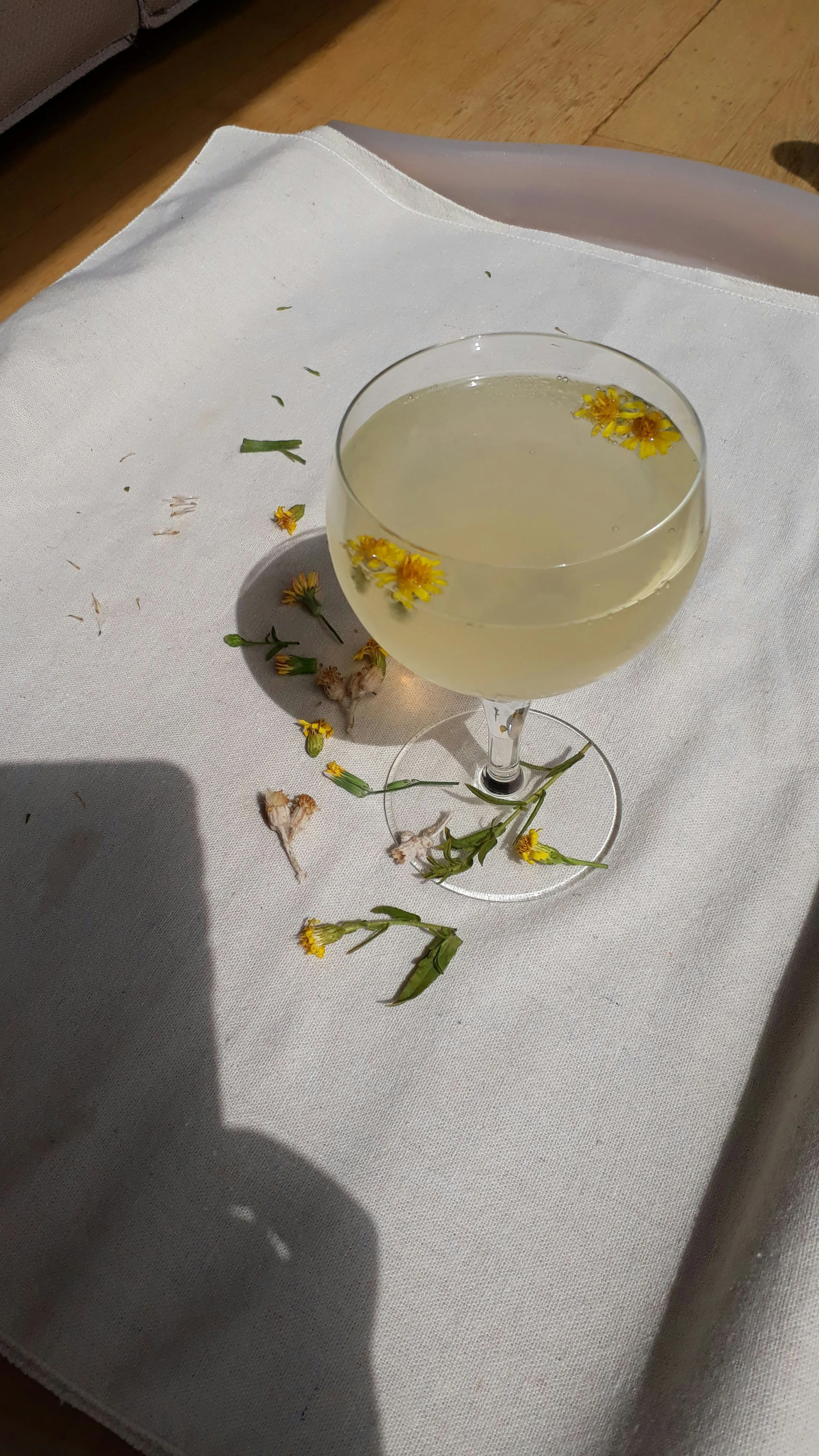 A glass of white wine with yellow flowers floating on top, placed on a white cloth with scattered yellow and green flower petals and stems.