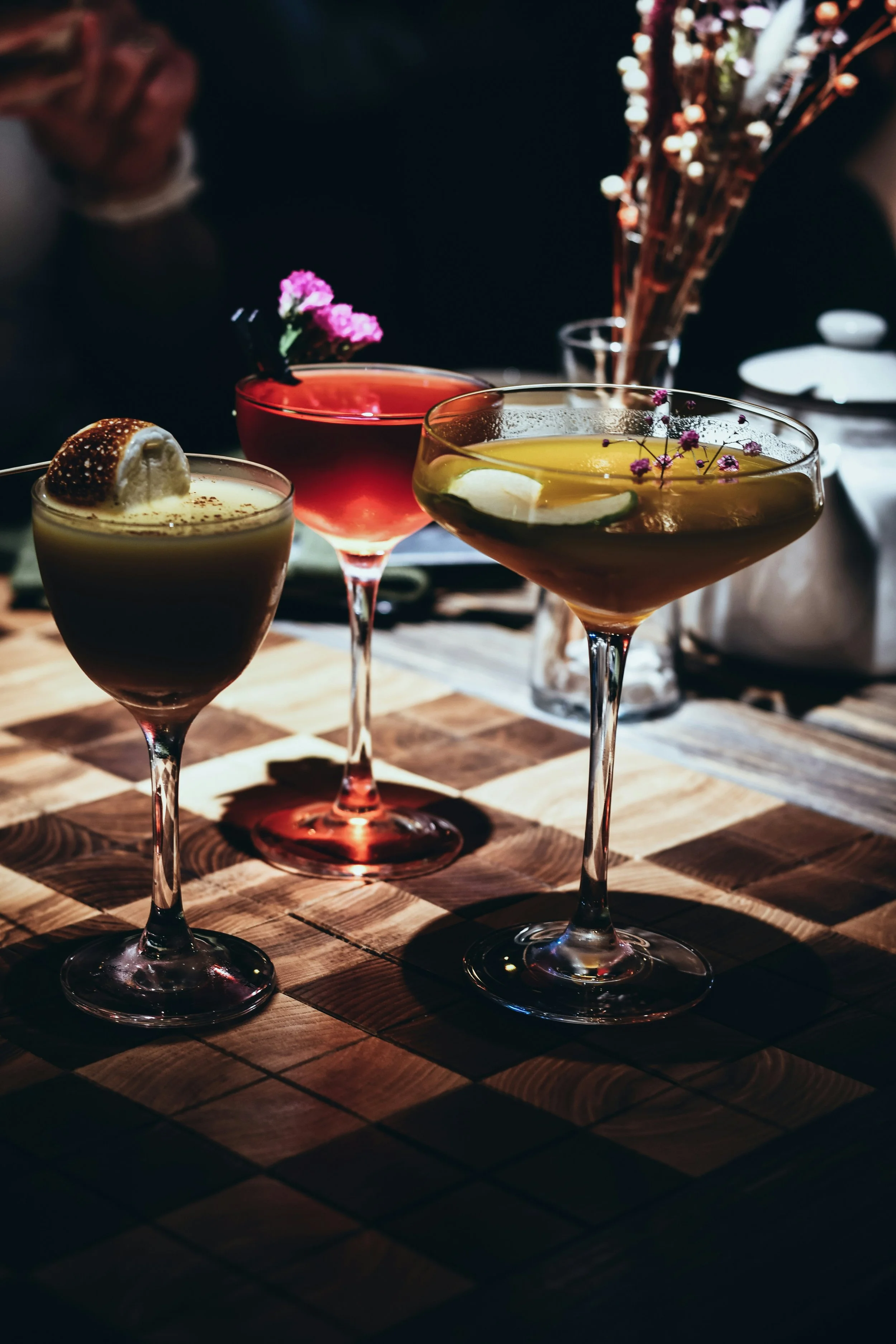 Three cocktails with garnishes on a wooden table, background with blurred flowers in a vase, dim lighting.
