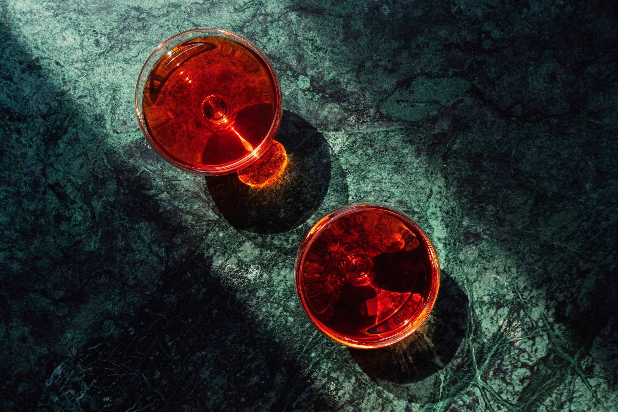 Two glasses of red wine placed on a dark textured surface, viewed from above, with sunlight reflections creating patterns on the liquid.