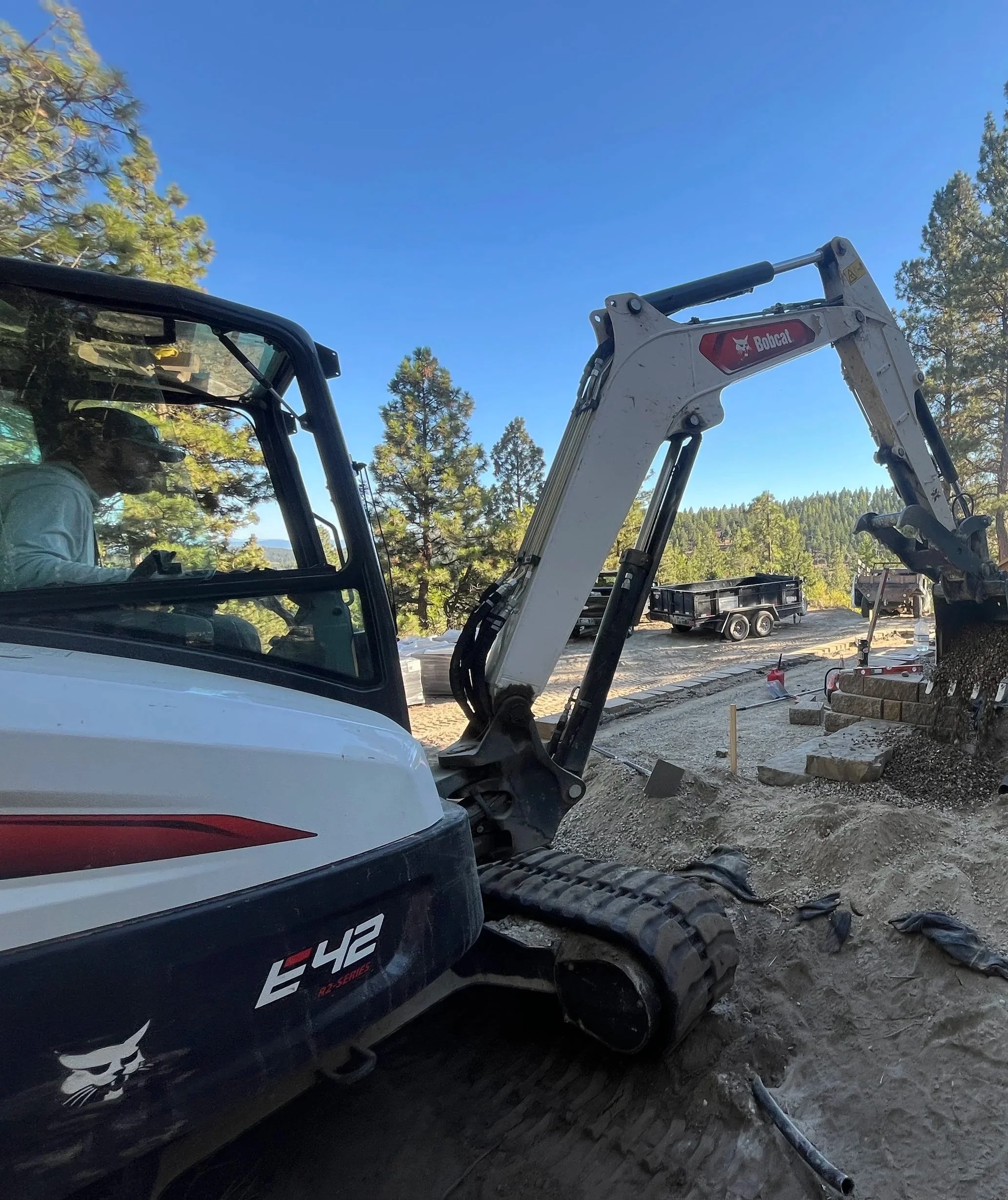 Construction site with a Bobcat mini excavator and a worker operating it, surrounded by trees and construction materials.