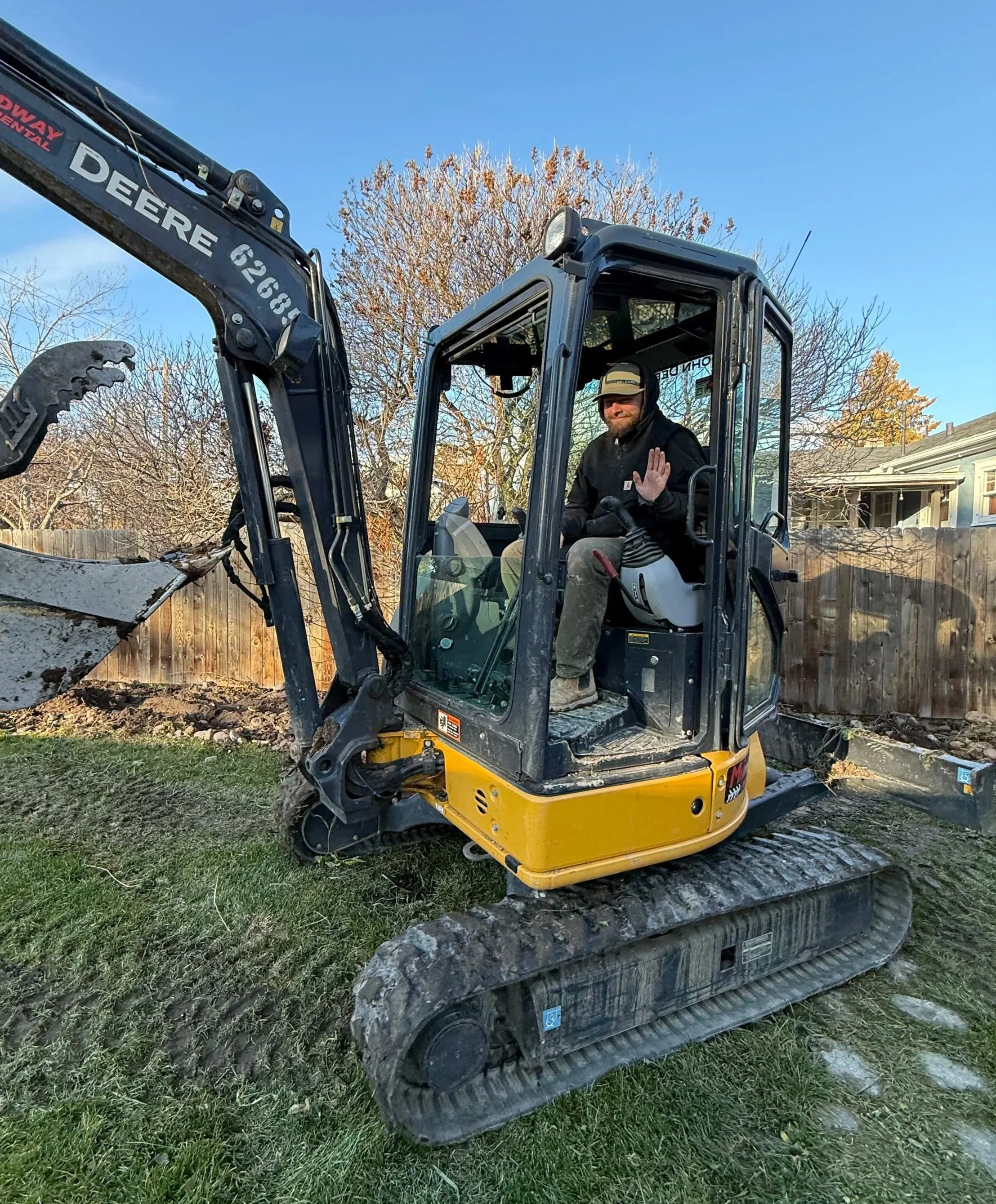 A man sitting in the operator's seat of a yellow and black mini excavator, waving at the camera, in a backyard with trees and a wooden fence in the background.