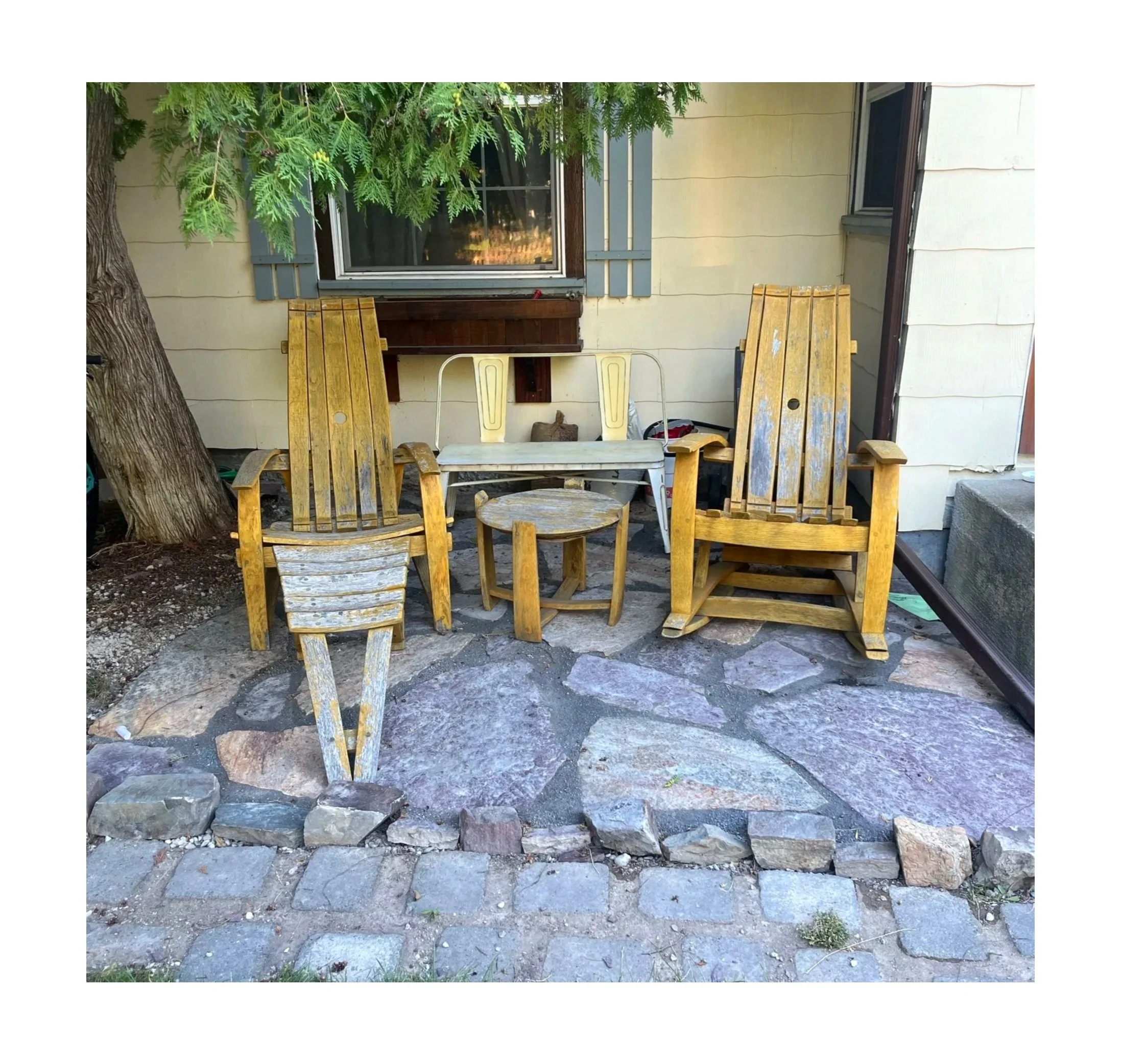 Outdoor patio with weathered wooden chairs, small table, stone paving, tree, house wall, window, and various objects in the background.