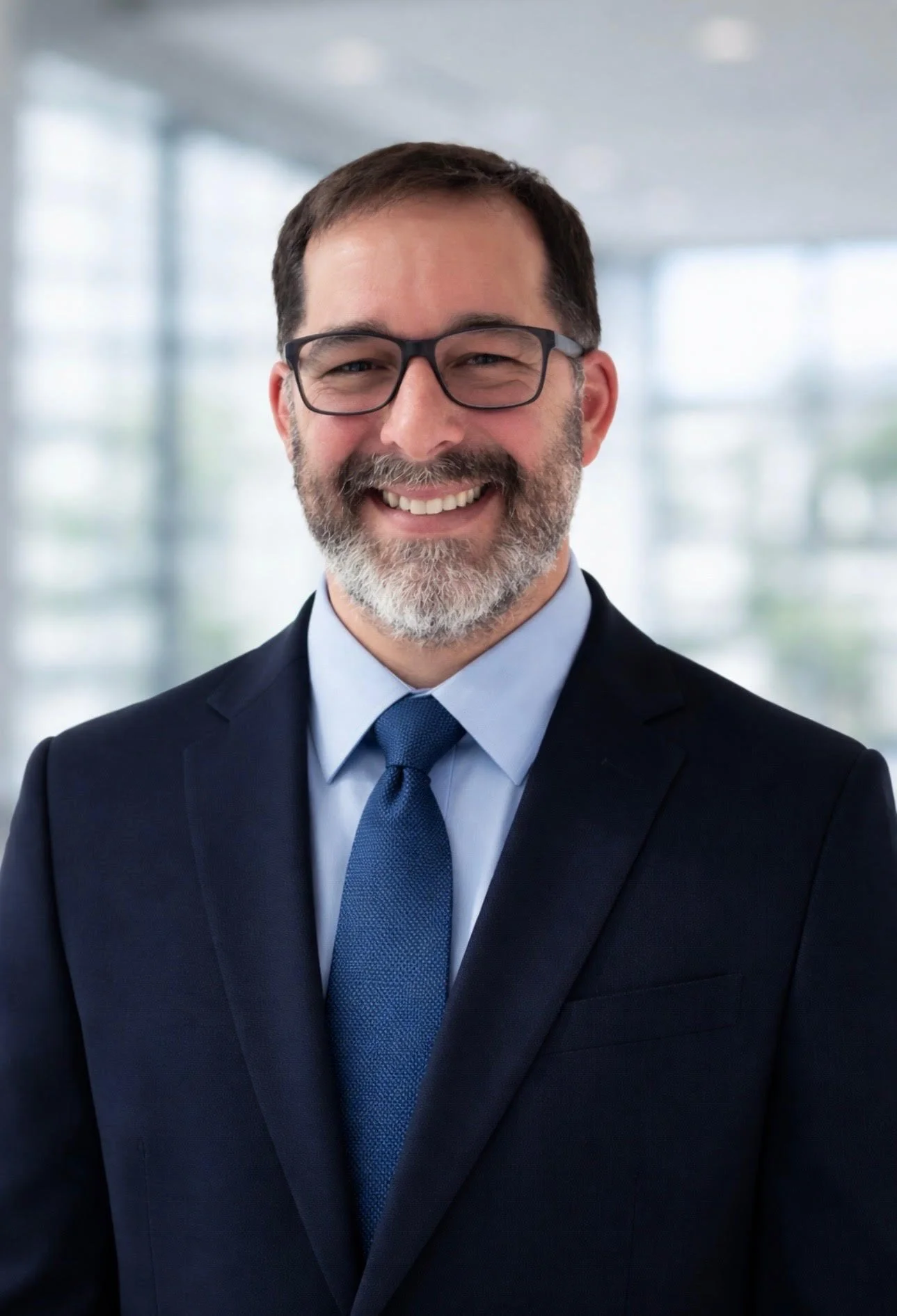 A smiling man with glasses, a beard, wearing a dark suit, light blue shirt, and matching blue tie, standing in a modern office with large windows in the background.