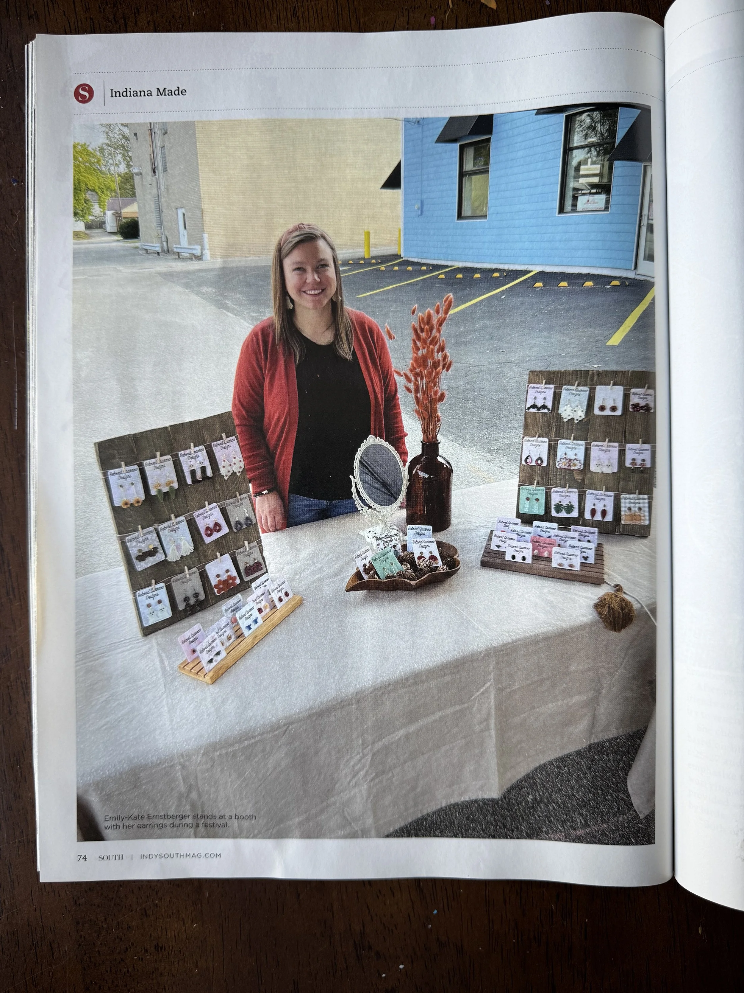 A woman standing behind a jewelry booth with various earrings displayed on racks, a brown vase with dried orange flowers, and a small ornate mirror, outdoors in a parking lot next to a blue building.
