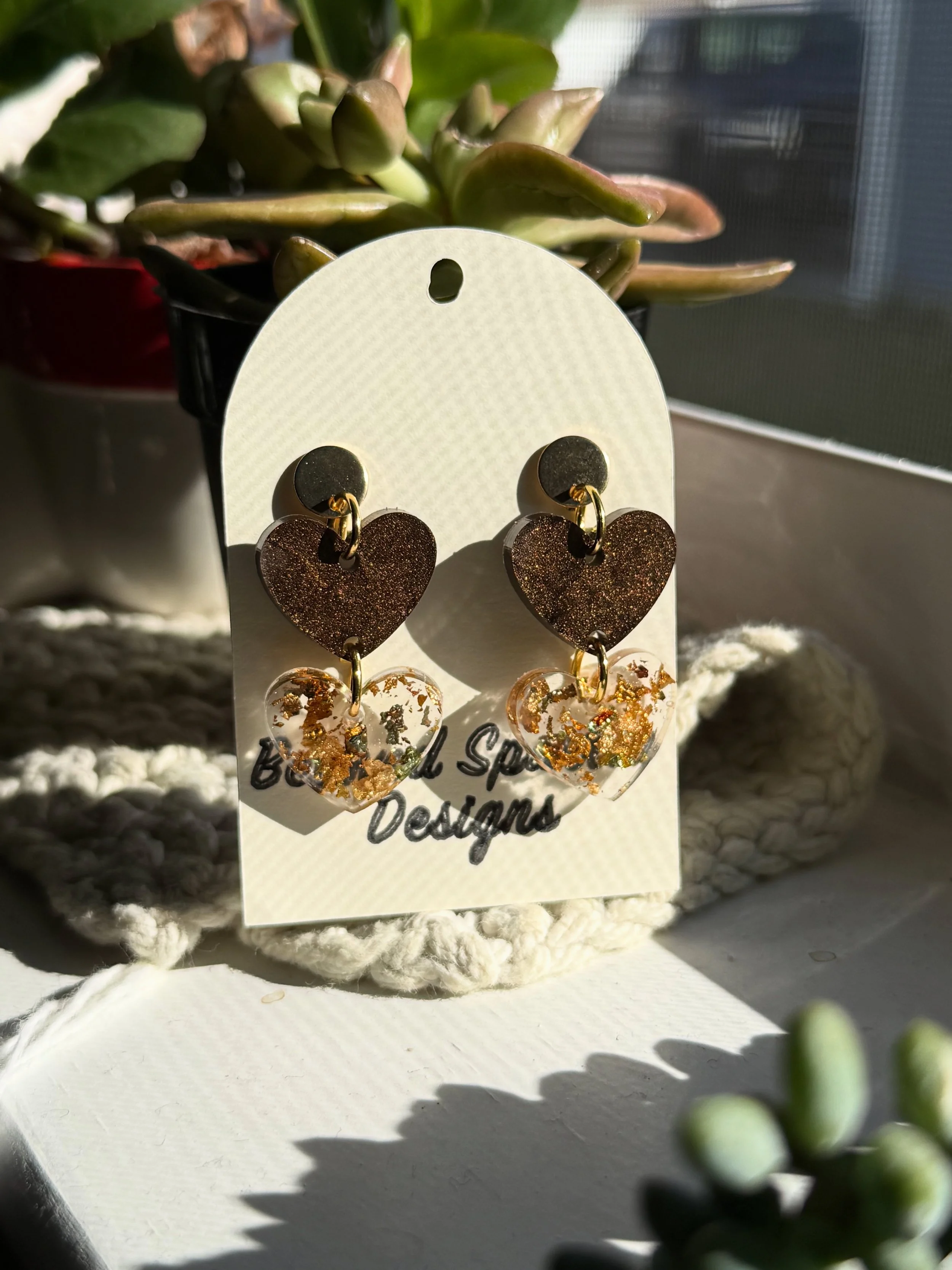 A pair of glittery gold heart-shaped earrings on a white display card with text. The earrings are in front of a potted succulent plant with thick green leaves. Sunlight casts shadows on the surface.