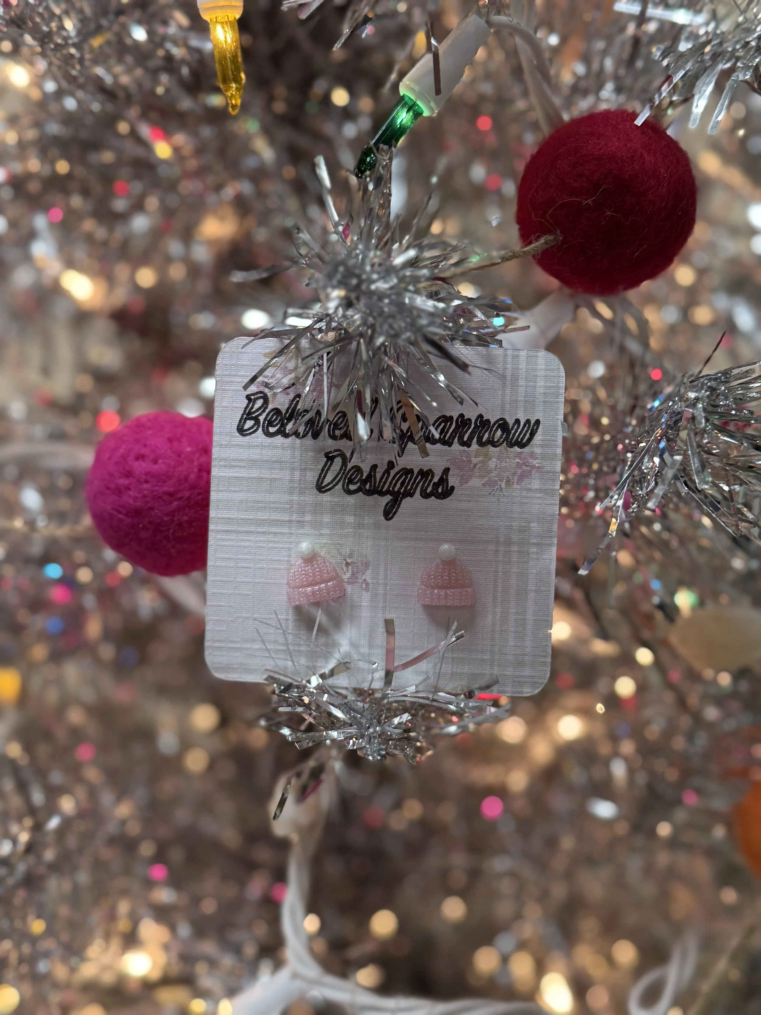 Close-up of a Christmas tree ornament with pink felt earrings and a red felt ball on a silver tinsel-covered tree.