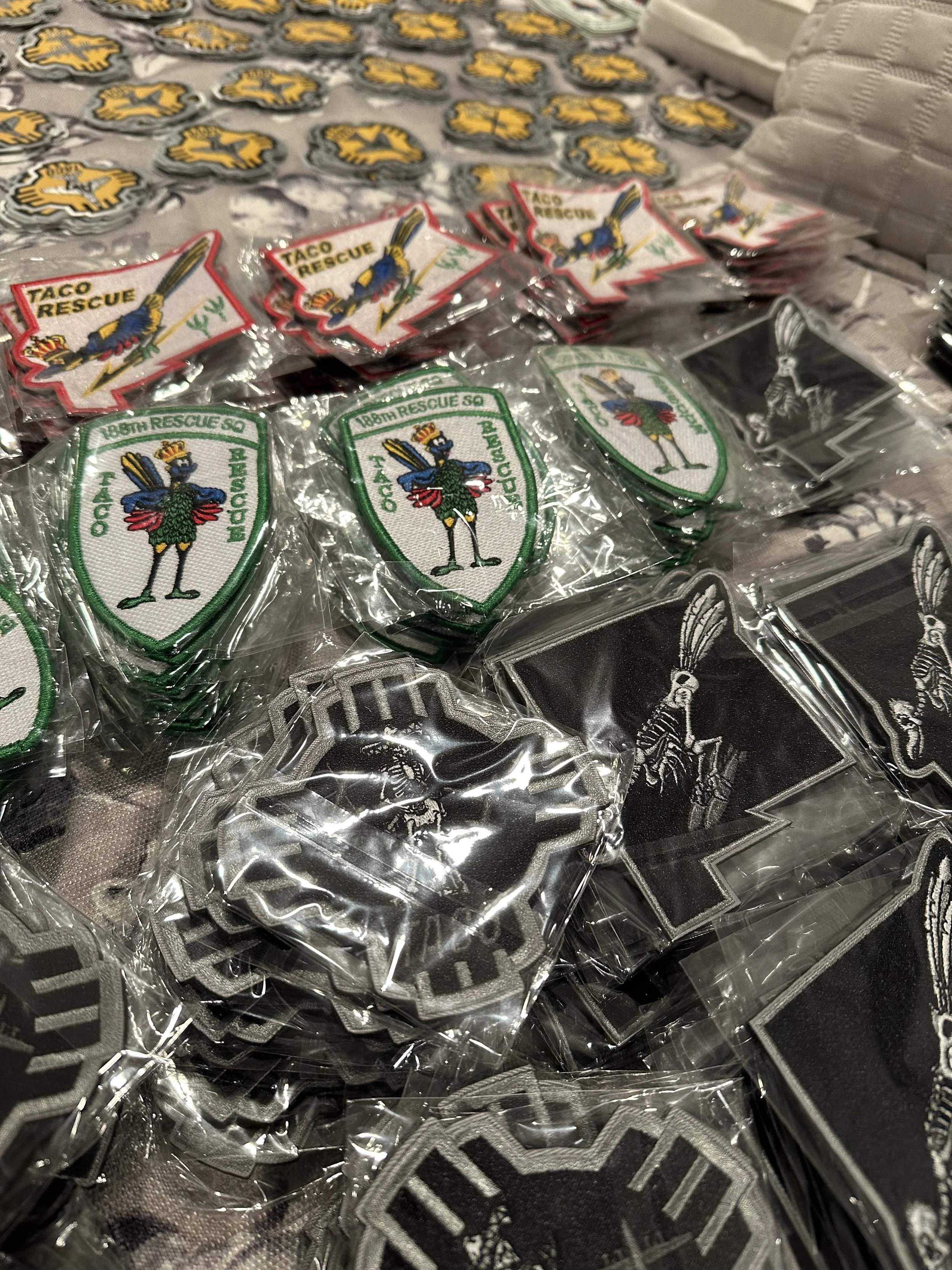 A collection of embroidered patches, including Taco Rescue patches with a cartoon roadrunner, TACO Rescue patches featuring a roadrunner with a crown, and black patches with a skeleton figure in a sombrero, all packaged in clear plastic.