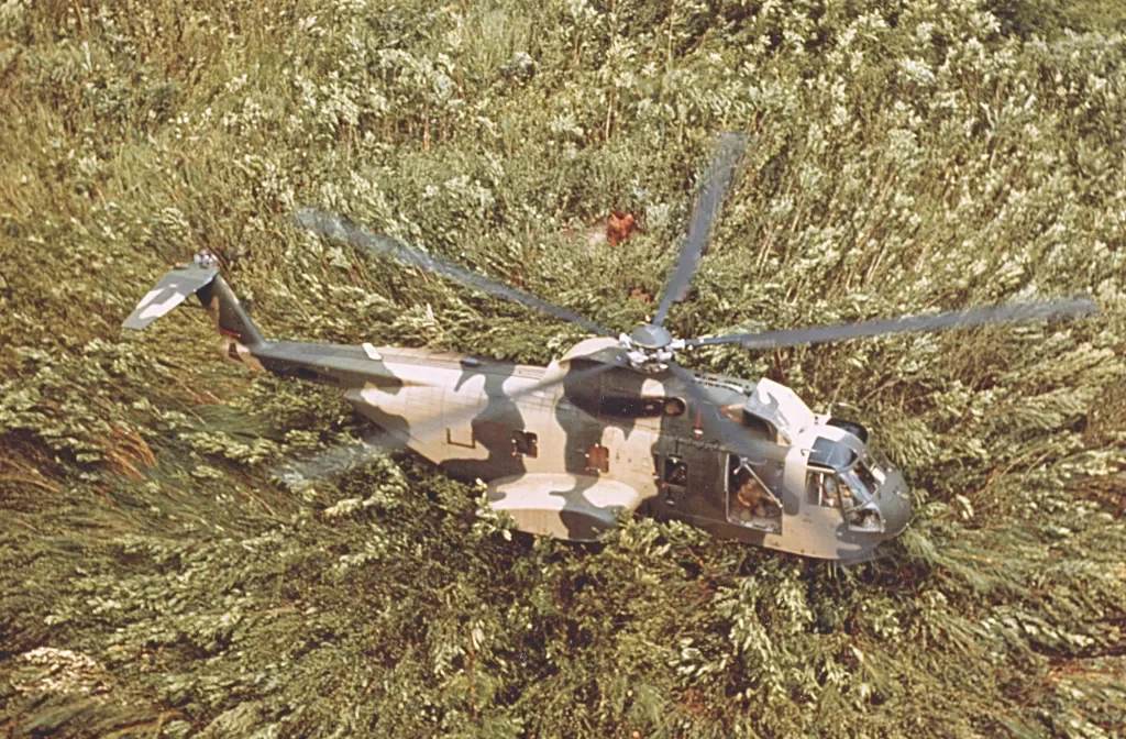 A HH-3E Jolly Green Giant helicopter lands in a forested area with dense green foliage.