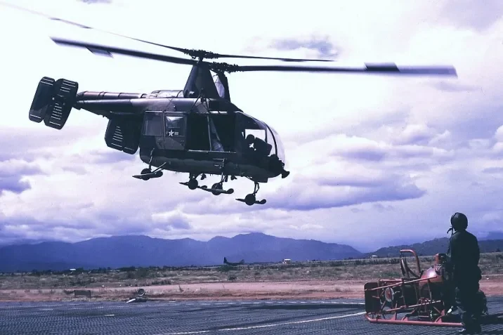 A HH-43B Huskie helicopter flying above a runway with a person standing next to equipment.