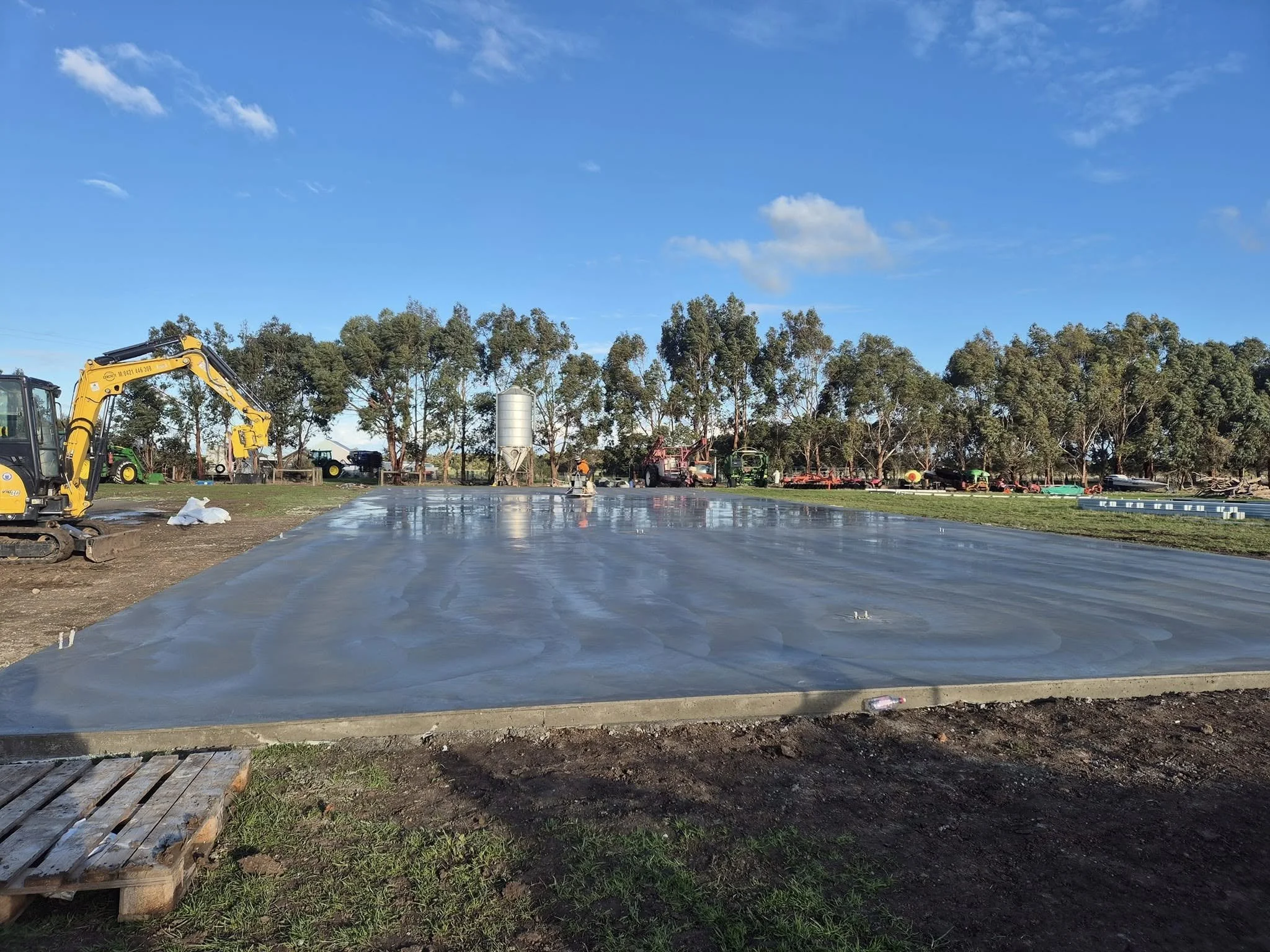 A construction site with a large freshly poured concrete slab, a yellow excavator on the left, and machinery and trees in the background under a blue sky with some clouds.