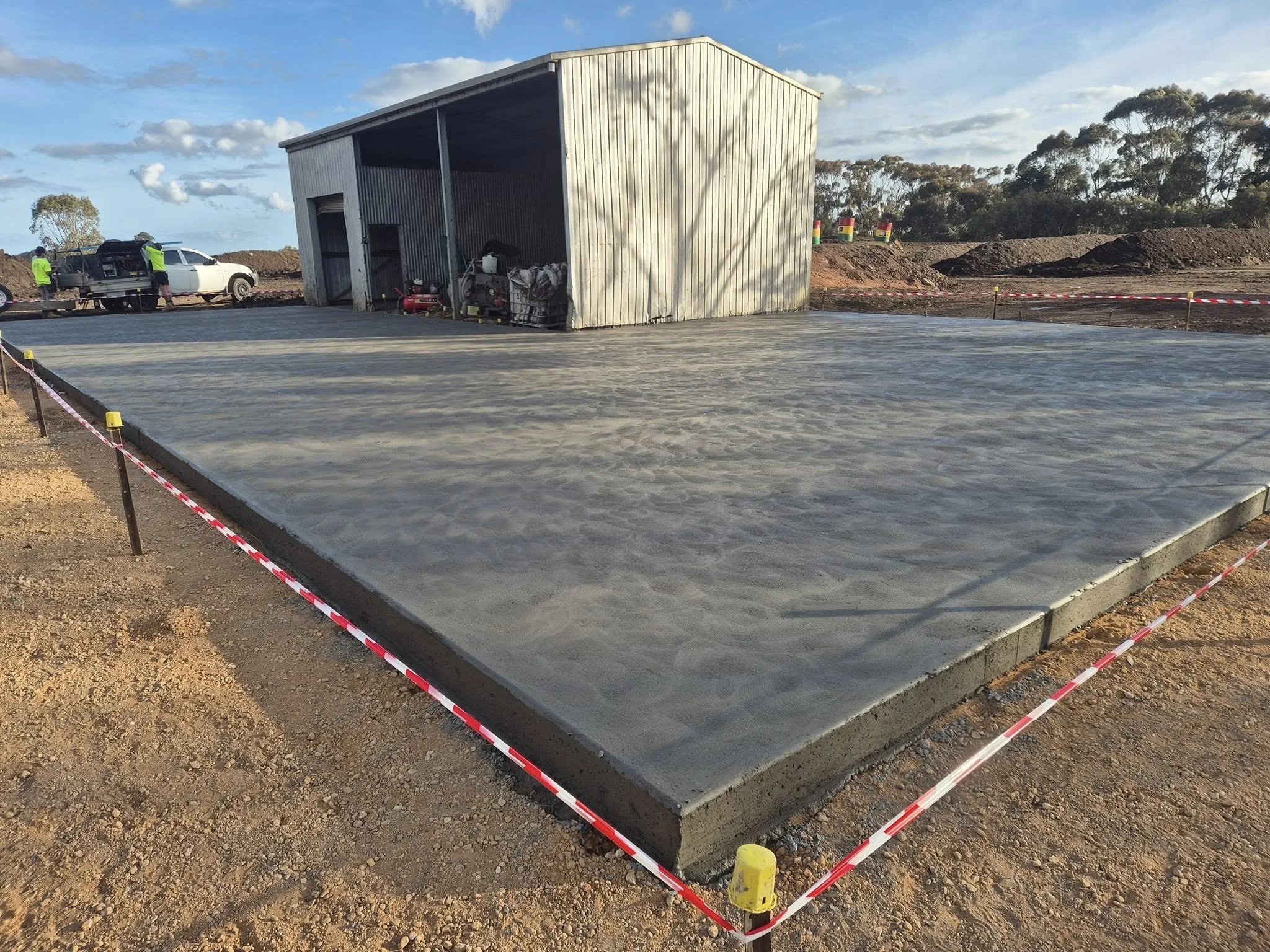Freshly poured concrete slab on construction site with a small workshop building in background, surrounded by safety tape and construction vehicles.