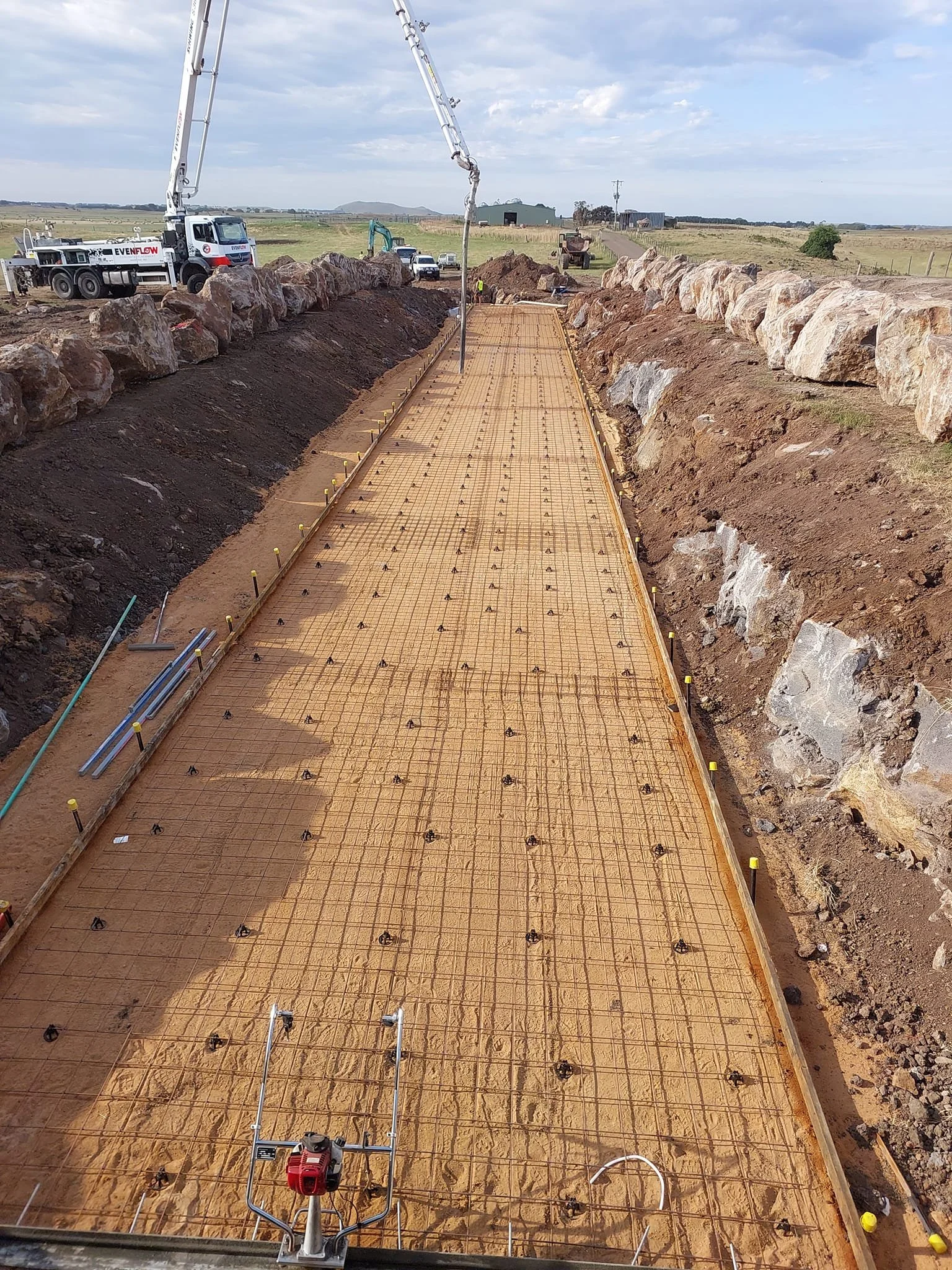 Construction site with a reinforced concrete foundation being prepared, with rebar grid and orange safety barriers, surrounded by dirt and rocks, under a partly cloudy sky.