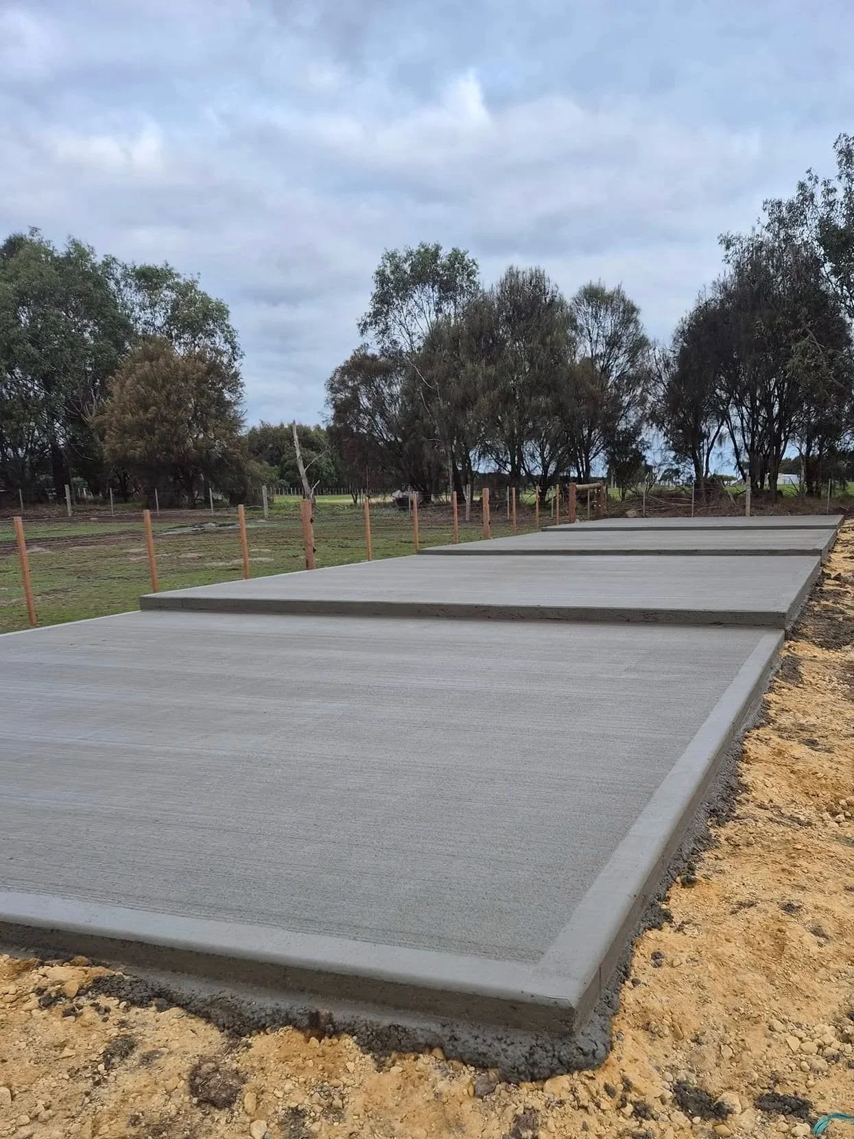 Newly poured concrete sidewalk sections on a construction site with trees and overcast sky in the background.