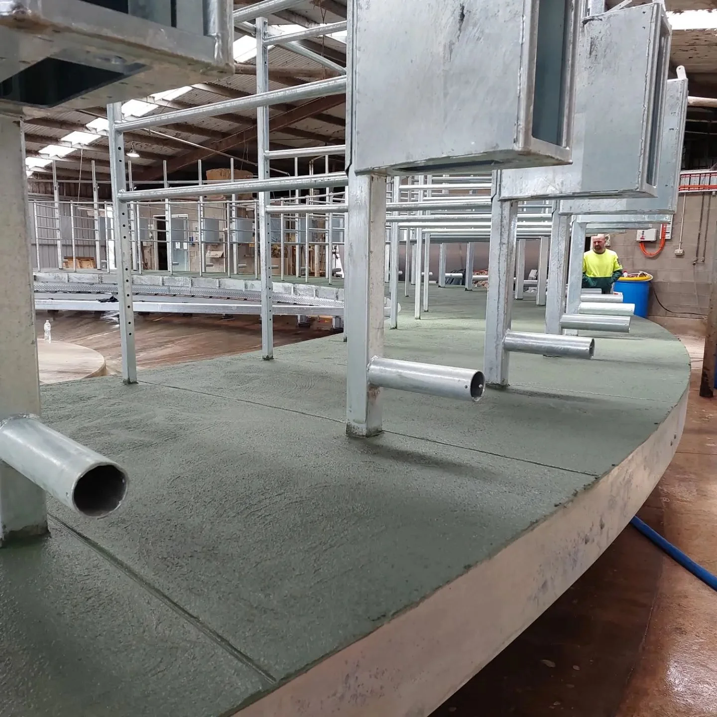 Metal framework being assembled on a concrete surface inside a warehouse, with a worker in a neon yellow safety vest standing in the background.