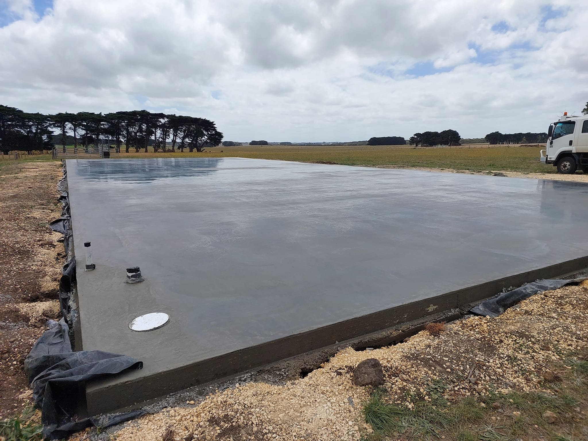Freshly poured concrete slab on a construction site with a truck parked in the background and trees on the horizon under cloudy skies.