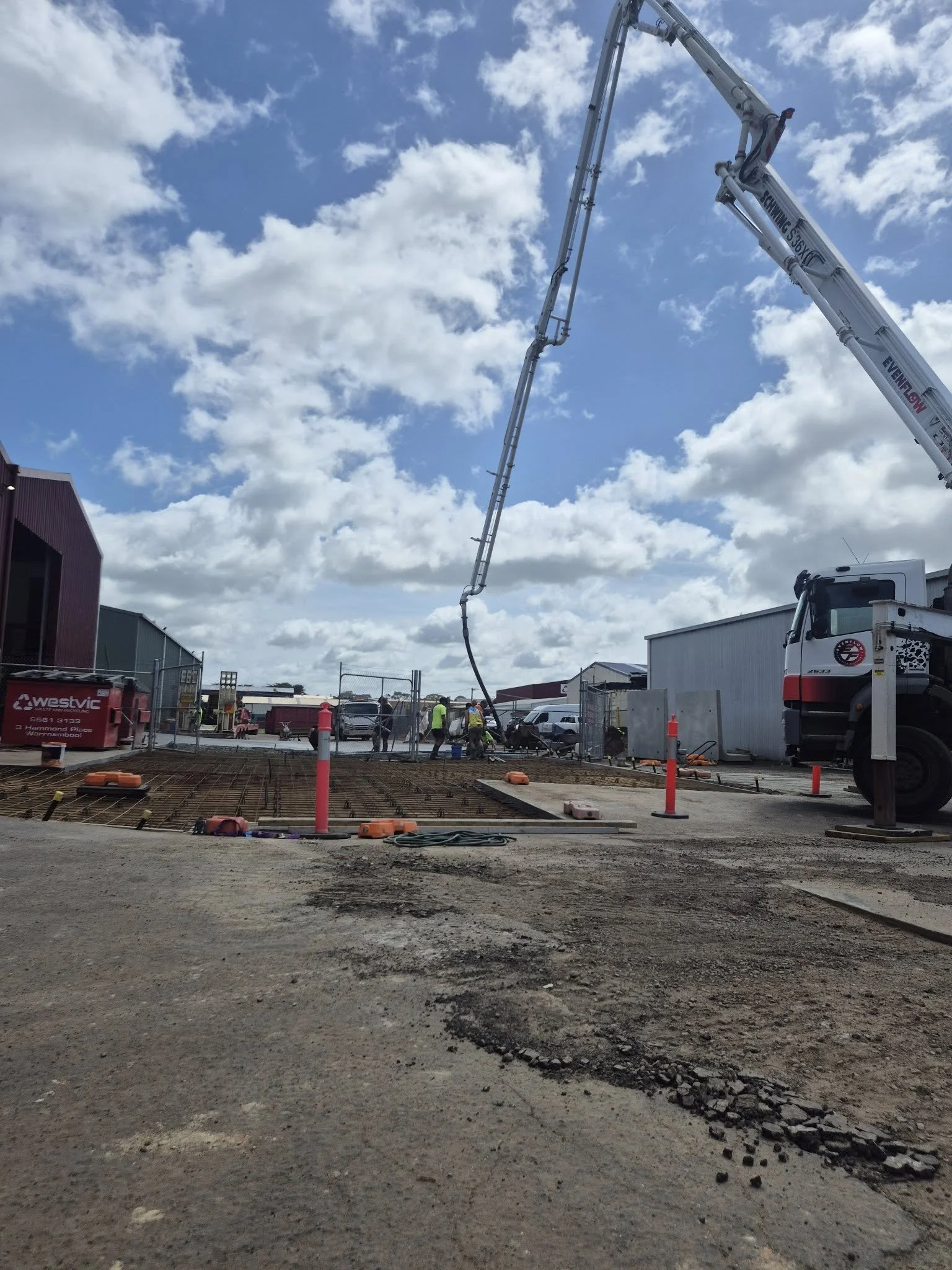 Construction site with concrete pouring equipment and workers, surrounded by fencing and buildings, under a partly cloudy sky.
