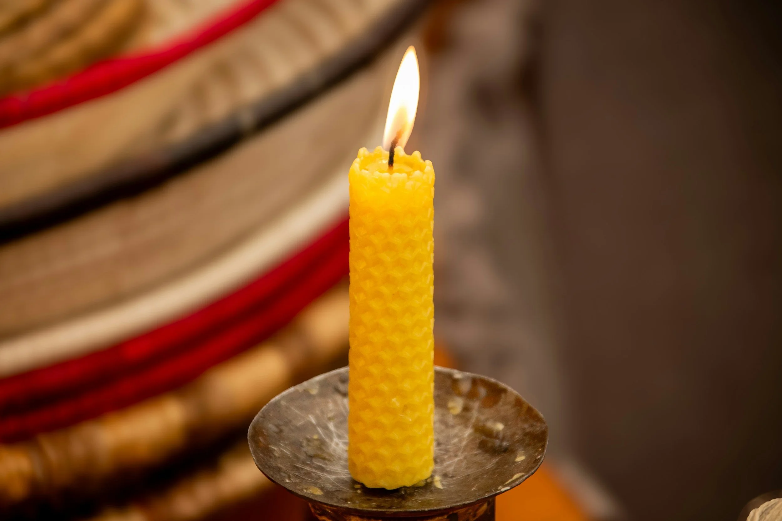 A tall yellow beeswax candle with a flame burning on top, placed on a metal candle holder.